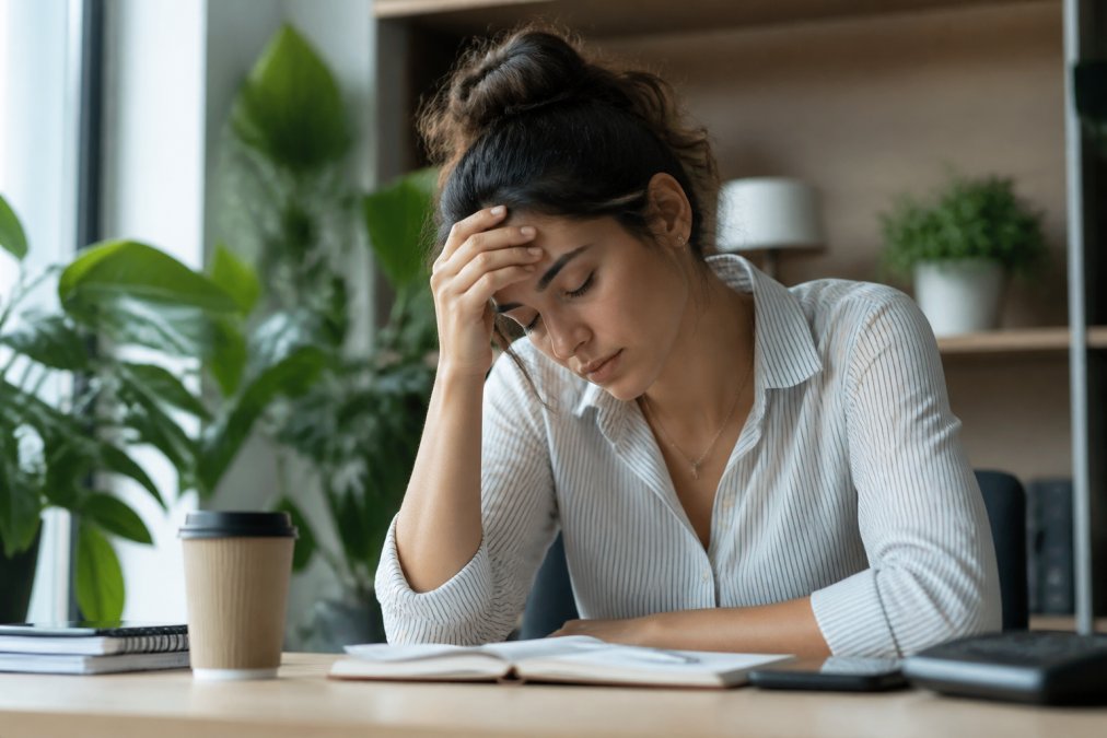 Woman experiencing fatigue and stress, sitting at desk with hand on forehead, showing signs of physical exhaustion and frustration in natural office lighting