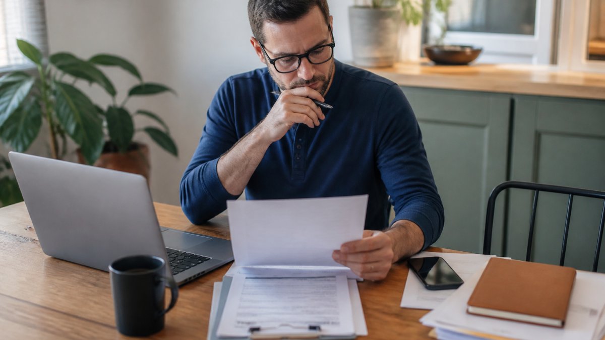 A patient sitting at a desk with a laptop, reviewing a prior authorization form and medical documents, with a phone nearby for calling the insurance company